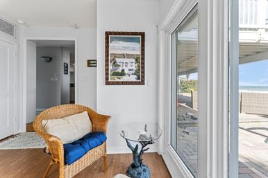 Oceanfront sitting nook with wicker armchair, blue cushion and beige pillow, glass side table with sculpted dolphin base, framed beach print on white wall and sliding glass door opening to a patio and sandy shoreline