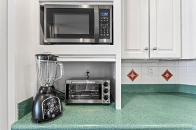 Green laminate kitchen countertop with built-in stainless steel microwave, stainless toaster oven and black blender under white cabinets and tiled backsplash with red diamond accents.