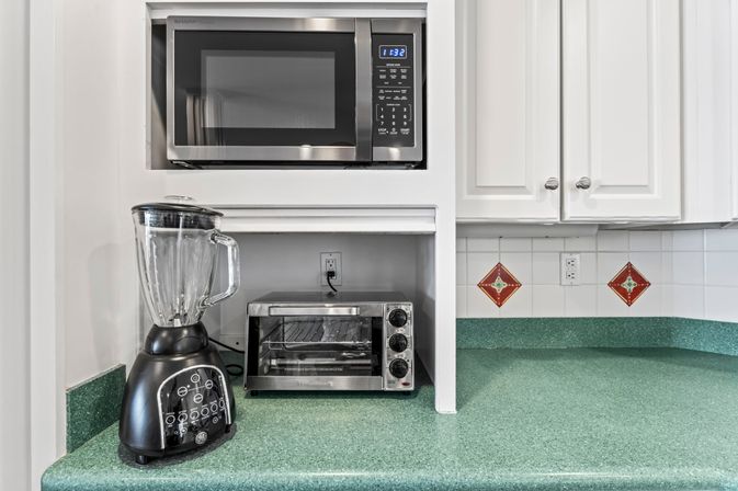 Green laminate kitchen countertop with built-in stainless steel microwave, stainless toaster oven and black blender under white cabinets and tiled backsplash with red diamond accents.