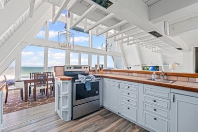 Sunlit oceanfront A-frame kitchen with white vaulted beams, wood countertops, stainless steel range, dining area, and panoramic windows framing a sandy beach and blue sea.