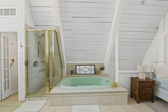 Cozy attic-style bathroom with white shiplap beamed ceiling, mint-green jetted soaking tub with brass faucet, glass shower with brass trim, tiled surround and wooden nightstand beside a bedroom nook.