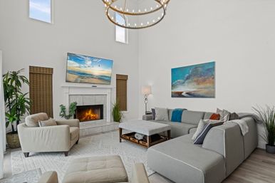 Bright two-story modern living room with high ceiling and round chandelier, cozy fireplace under a wall-mounted TV, gray sectional sofa, armchair and ottoman, coffee table, abstract wall art and indoor plants.