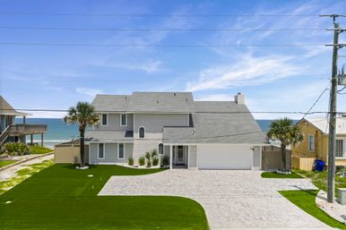 Sunny coastal two-story gray beach house with garage, brick driveway, palm trees and manicured lawn overlooking the turquoise ocean and clear blue sky.