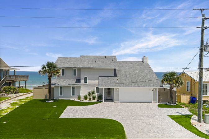 Sunny coastal two-story gray beach house with garage, brick driveway, palm trees and manicured lawn overlooking the turquoise ocean and clear blue sky.