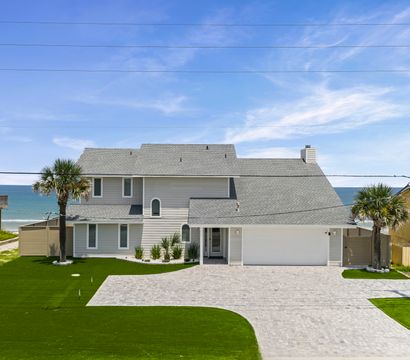 Sunny coastal two-story gray beach house with garage, brick driveway, palm trees and manicured lawn overlooking the turquoise ocean and clear blue sky.