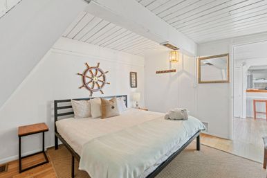 Bright nautical-themed attic bedroom with white shiplap walls, ship’s wheel wall decor, king bed with neutral linens, hardwood floors and folded towels