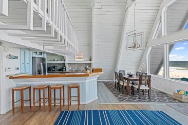Sunlit A-frame beachfront interior with white vaulted wood ceiling, coastal kitchen and wooden breakfast bar with four stools, dining table for six, and large windows framing sandy beach and ocean views.