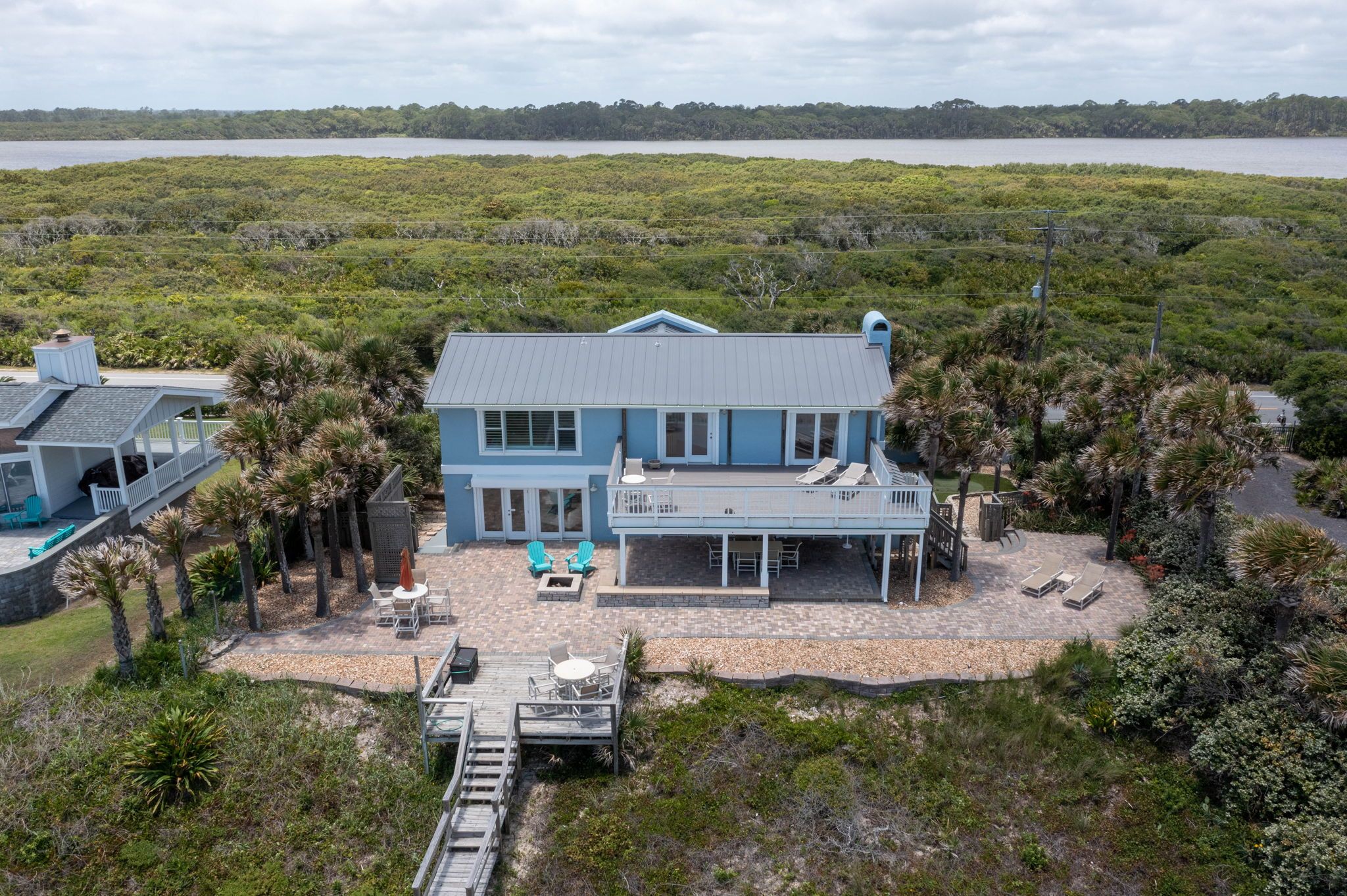 Blue two-story coastal beach house with metal roof, spacious second-story deck and patio with lounge chairs, fire pit and a wooden boardwalk over dunes leading to the shore, surrounded by palm trees and coastal marsh under a cloudy sky.