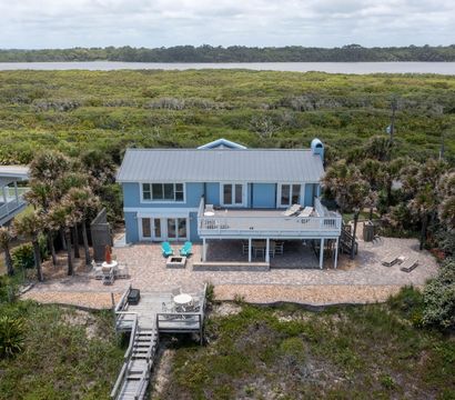 Blue two-story coastal beach house with metal roof, spacious second-story deck and patio with lounge chairs, fire pit and a wooden boardwalk over dunes leading to the shore, surrounded by palm trees and coastal marsh under a cloudy sky.