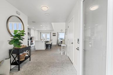 Bright coastal open-plan entryway leading to a white kitchen with island and bar stools, round wall mirror, potted fiddle-leaf fig, hardwood floors and distant ocean view through sliding doors.