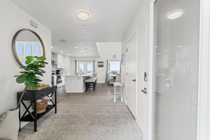 Bright coastal open-plan entryway leading to a white kitchen with island and bar stools, round wall mirror, potted fiddle-leaf fig, hardwood floors and distant ocean view through sliding doors.