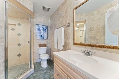 Cozy residential bathroom with glass-enclosed shower and diamond-tile accents, white vanity and sink, gold-framed mirror, toilet, towel rack and abstract wall art.