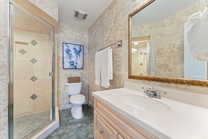 Cozy residential bathroom with glass-enclosed shower and diamond-tile accents, white vanity and sink, gold-framed mirror, toilet, towel rack and abstract wall art.