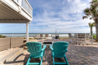Beachfront patio with two turquoise Adirondack chairs facing a built-in fire pit and boardwalk to the ocean, brick pavers, palm trees and outdoor dining set under a blue sky.