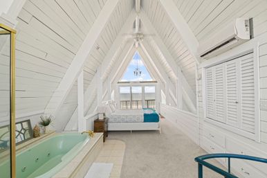 Bright white A-frame loft bedroom with shiplap walls, mint soaking tub, bed and balcony overlooking beachfront ocean views