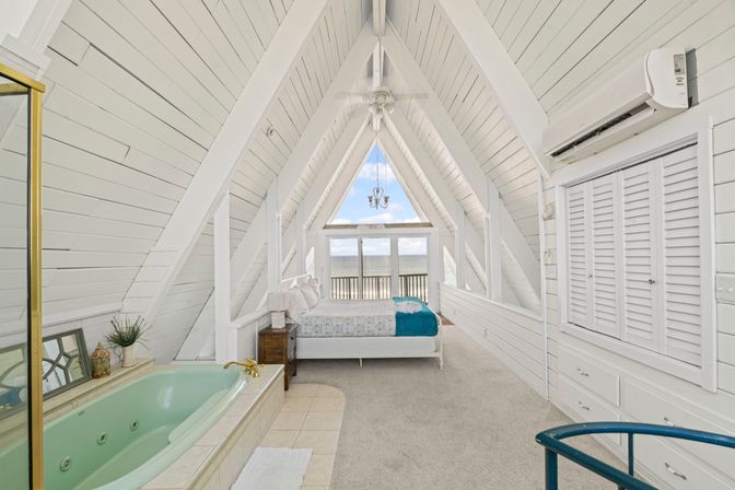 Bright white A-frame loft bedroom with shiplap walls, mint soaking tub, bed and balcony overlooking beachfront ocean views