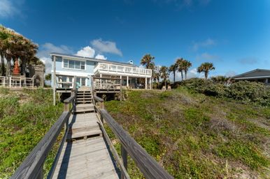 Oceanfront beach house on sand dunes with a wooden boardwalk and stairs, palm trees and bright blue sky.