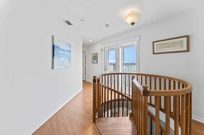 Sunny coastal upstairs landing with curved wooden spiral staircase, light oak floors, white walls, framed art and French doors opening to a balcony with ocean and sky view.