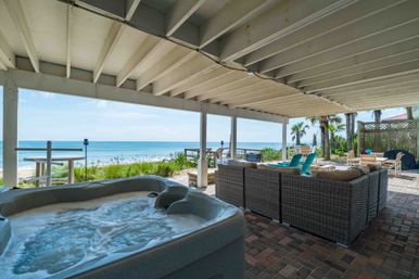 Covered beachfront patio with bubbling hot tub in foreground, wicker sectional sofa, turquoise Adirondack chairs, palm trees and unobstructed ocean view under a white wooden beam ceiling.