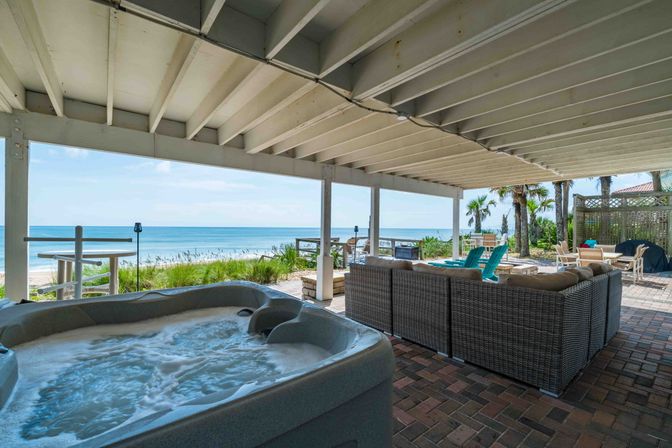 Covered beachfront patio with bubbling hot tub in foreground, wicker sectional sofa, turquoise Adirondack chairs, palm trees and unobstructed ocean view under a white wooden beam ceiling.