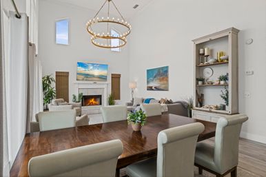 Bright open-concept living and dining room with high ceilings and double-ring chandelier, wooden dining table with upholstered chairs in foreground, cozy seating area with lit fireplace and wall-mounted TV, neutral decor, tall windows, shelving and potted plants