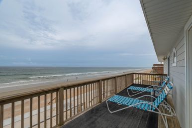 Oceanfront balcony with wooden railing and three turquoise woven lounge chairs overlooking a wide sandy beach and gentle waves under a cloudy sky.