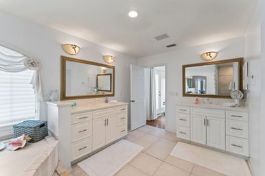 Bright spa-like master bathroom with double white vanities and gold-framed mirrors, wall sconces, tiled floor, built-in tub ledge with wicker basket and seashells, open doorway to bedroom.