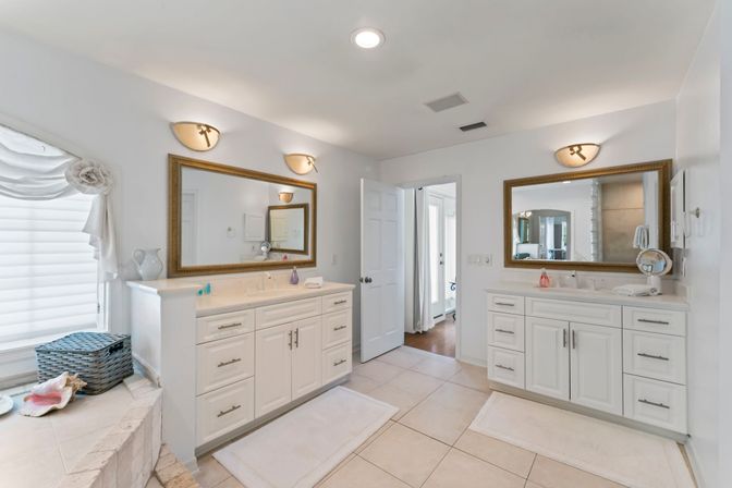 Bright spa-like master bathroom with double white vanities and gold-framed mirrors, wall sconces, tiled floor, built-in tub ledge with wicker basket and seashells, open doorway to bedroom.