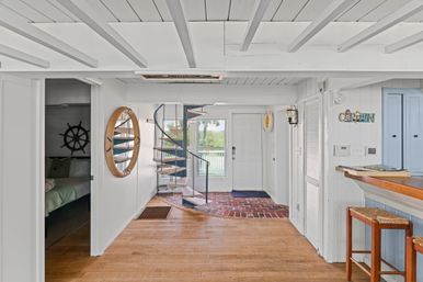 Bright coastal cottage entryway with white paneled walls and exposed beams, wood floor, black spiral staircase, rattan porthole mirror, nautical accents, bar stools, and a glass door to a waterfront balcony.