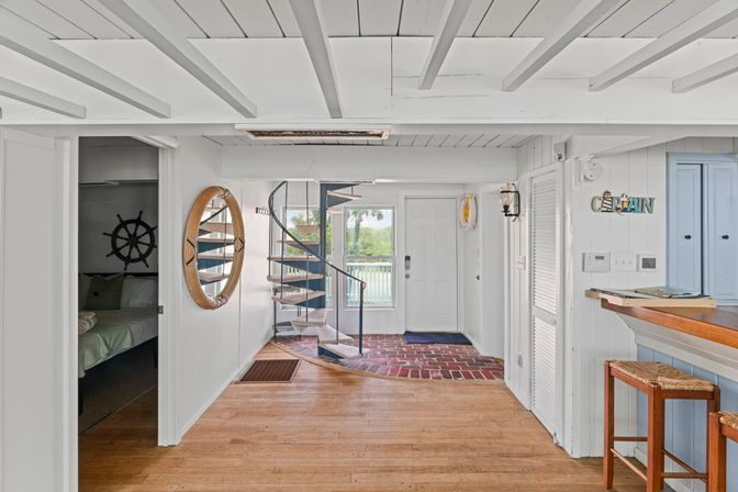 Bright coastal cottage entryway with white paneled walls and exposed beams, wood floor, black spiral staircase, rattan porthole mirror, nautical accents, bar stools, and a glass door to a waterfront balcony.