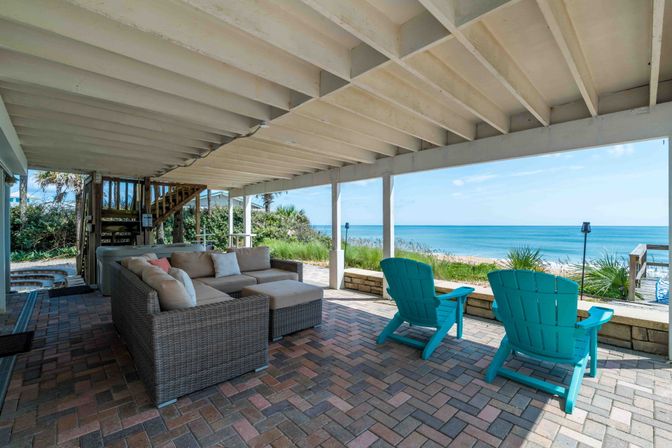 Oceanfront covered patio under a raised deck with brick pavers, wicker sectional sofa and turquoise Adirondack chairs facing dunes and a bright blue ocean on a sunny day