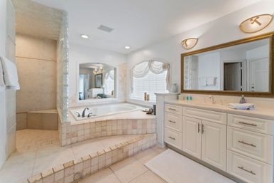 Sunlit spa-like master bathroom with raised tiled soaking tub, glass-block walk-in shower with bench, long white double-sink vanity and large framed mirror in neutral tile