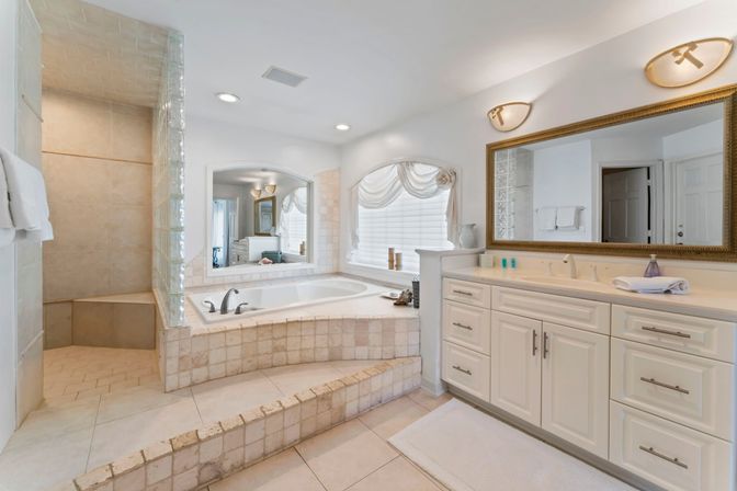 Sunlit spa-like master bathroom with raised tiled soaking tub, glass-block walk-in shower with bench, long white double-sink vanity and large framed mirror in neutral tile