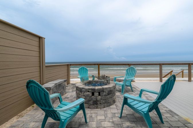 Oceanfront deck with a circular stone fire pit and four teal Adirondack chairs overlooking a sandy beach and cloudy sea.