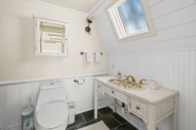 Cozy attic bathroom with skylight, white beadboard walls, wicker vanity featuring a floral sink and brass faucet, and a modern white toilet.