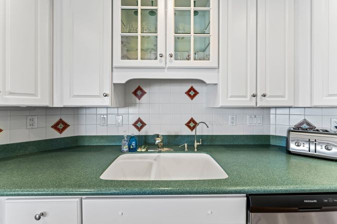 Bright modern kitchen sink with white cabinets and glass-front upper cabinet, green speckled countertop, white double-basin sink and chrome faucet, white tiled backsplash with red decorative tiles, stainless toaster and dishwasher, dish soap at the sink.