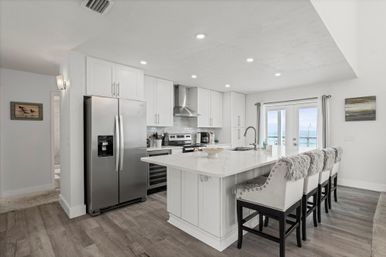 Bright modern coastal kitchen with white marble-look island, stainless steel appliances, four upholstered bar stools with faux-fur throws, and sliding glass doors framing an ocean view