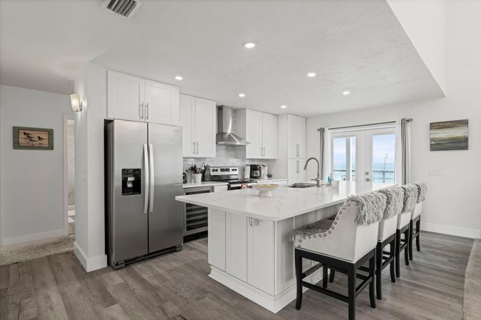 Bright modern coastal kitchen with white marble-look island, stainless steel appliances, four upholstered bar stools with faux-fur throws, and sliding glass doors framing an ocean view