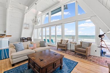 Bright A-frame beachfront living room with floor-to-ceiling windows and panoramic ocean view, cozy sofa, wicker chairs, wooden coffee table, chandelier, and a telescope overlooking a deck with blue Adirondack chairs.