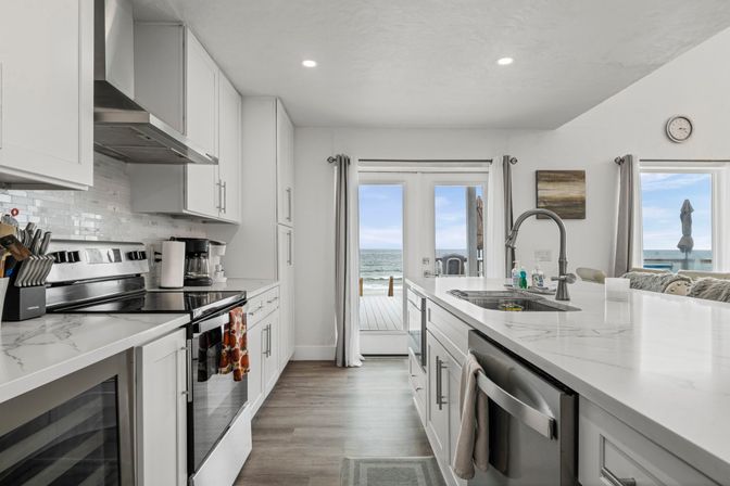 Bright modern white beachfront kitchen with marble-look countertops, stainless steel appliances, large island and sink, and French doors opening to an ocean-view deck.