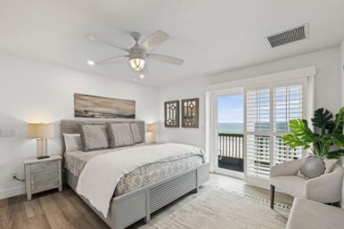 Sunlit coastal bedroom in a beachfront rental with a gray wood bed and layered neutral bedding, nightstands and lamps, ceiling fan, potted plant, and sliding glass doors with shutters opening to a balcony and ocean view