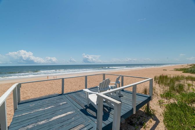 Sunny oceanfront beach scene with gentle waves and sandy shoreline, viewed from a weathered blue wooden deck with white railings and two white Adirondack chairs overlooking coastal dunes.