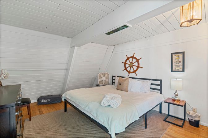 Coastal attic bedroom with white shiplap slanted ceiling, king bed with white linens and rolled towels, wooden ship’s wheel wall decor, bedside lamp, hardwood floors and dark dresser