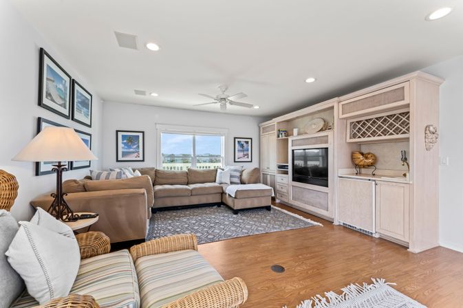 Sunny coastal living room with beige L-shaped sectional, wicker armchairs, whitewashed built-in entertainment center with TV and wine rack, hardwood floors, ceiling fan, framed beach prints, and a window overlooking a waterfront view.