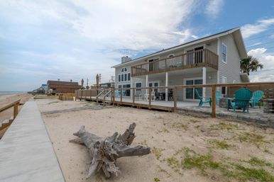 Oceanfront two-story beach house with upper balcony and ground-level deck, turquoise Adirondack chairs, a large driftwood log on the sandy shore and a concrete promenade under a cloudy blue sky