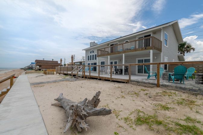 Oceanfront two-story beach house with upper balcony and ground-level deck, turquoise Adirondack chairs, a large driftwood log on the sandy shore and a concrete promenade under a cloudy blue sky