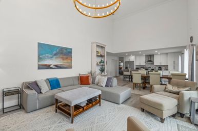 Open-plan modern living room and kitchen with vaulted ceiling, gray sectional sofa, ottoman coffee table, armchair and dining table under a circular brass chandelier.