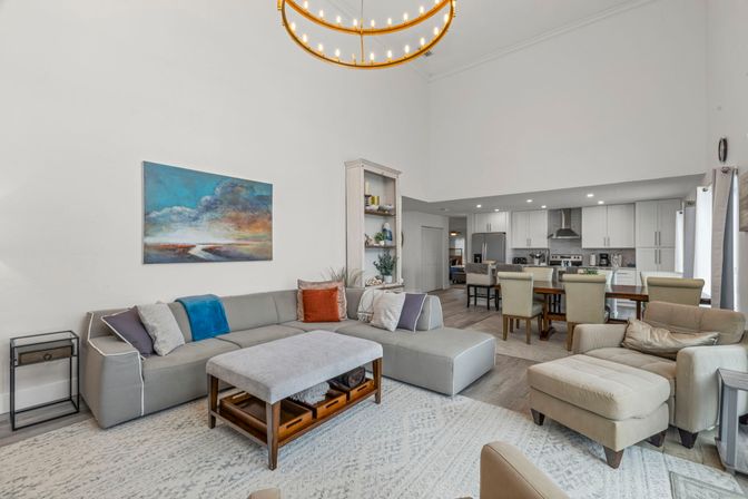 Open-plan modern living room and kitchen with vaulted ceiling, gray sectional sofa, ottoman coffee table, armchair and dining table under a circular brass chandelier.