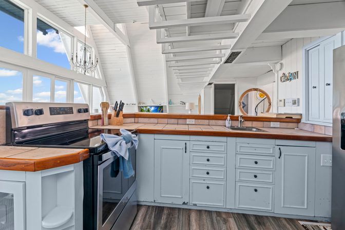 Sunlit coastal beach-cottage kitchen with vaulted white beam ceiling and large ocean-view windows, wood-trimmed tile counters, pale blue cabinets, stainless-steel stove, farmhouse sink and small chandelier.