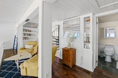 Bright coastal cottage loft with white shiplap walls and ceiling, cheerful yellow armchairs on a navy rug, built-in bookshelves with blue ladder, spiral staircase, wooden side table with lamp, and a small bathroom with skylight visible